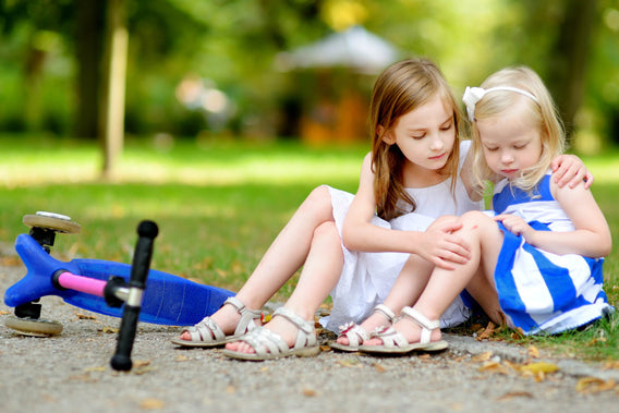 Two little girls sit on the ground; one cares for the other who has hurt her leg next to a scooter