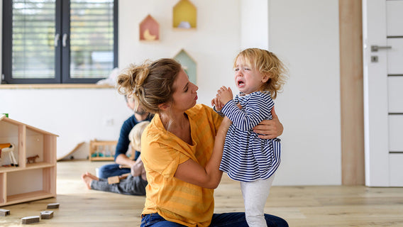 A woman kneels on the floor comforting a crying young child indoors, with toys and another adult and child in the background.