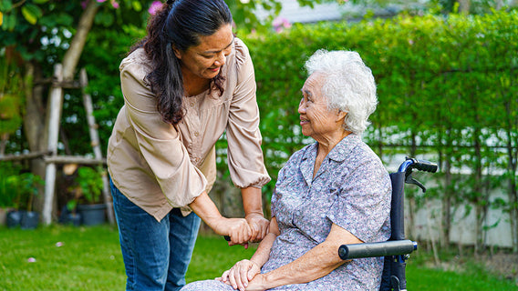 `A woman leans toward an elderly woman seated in a wheelchair outdoors, holding her hands and making eye contact in a moment of calm, attentive connection.`