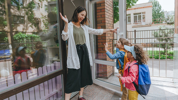 A teacher at a school entrance reaches out to greet two children arriving with backpacks.