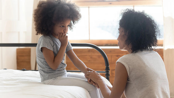 A worried child sits on a bed biting her finger while a supportive parent holds her hand, demonstrating secure attachment and connection during difficult moments in parenting.