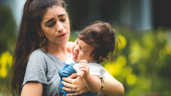 A mother holds her crying toddler outdoors, her face tense and overwhelmed.