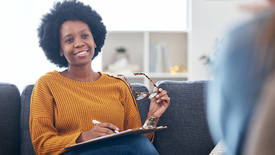 A therapist smiles warmly while seated on a couch, holding glasses and a clipboard. She exudes a friendly, approachable atmosphere.