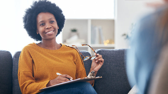 A therapist smiles warmly while seated on a couch, holding glasses and a clipboard. She exudes a friendly, approachable atmosphere.