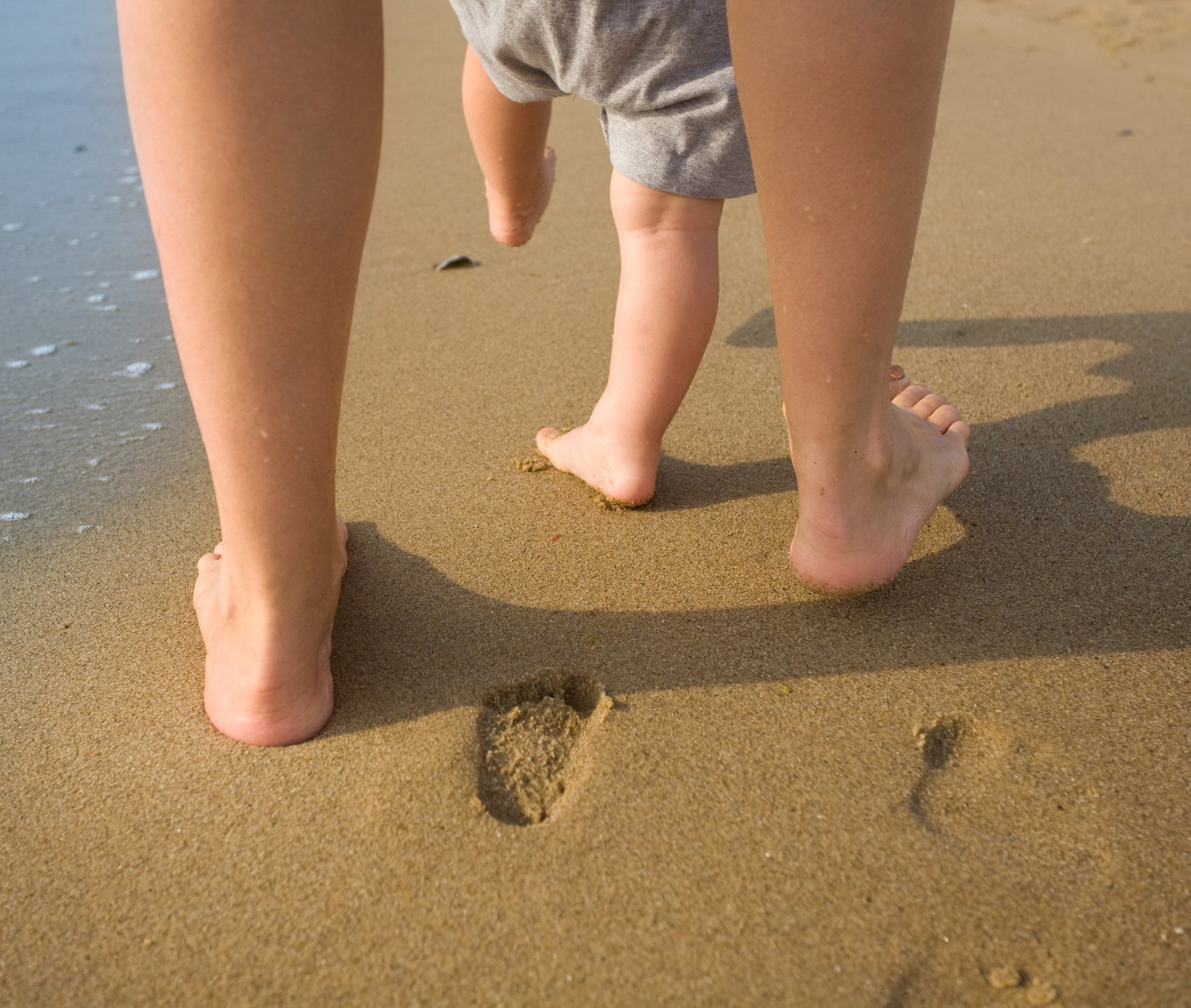 a mother's feet next to a toddler's feet as they walk along the beach leaving footprints together in the sand