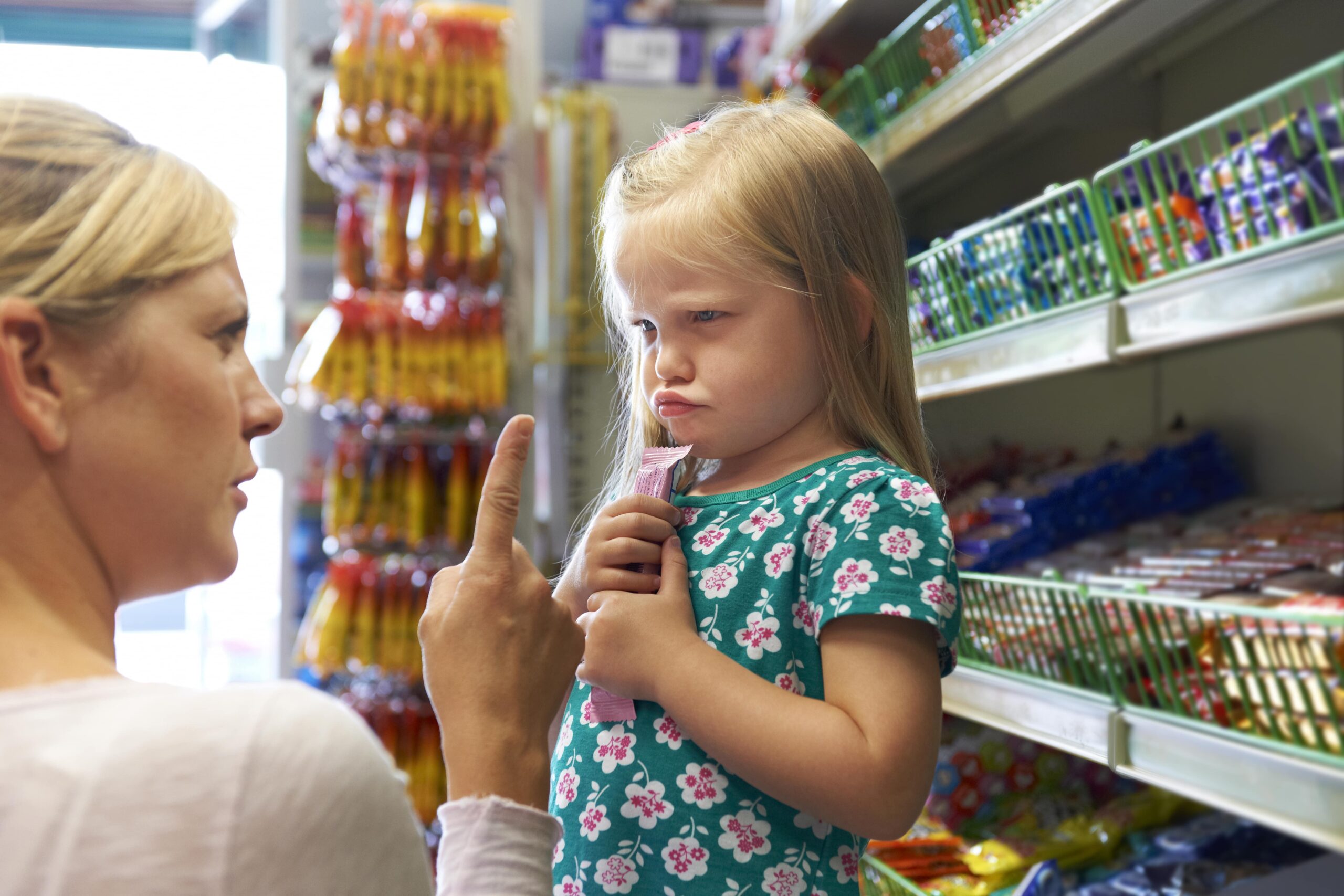a girl pouting at the grocery store as her mother tells her to put the candy bar back on the shelf